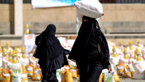 A woman in black carries a sack of grain on her head. There are lots of other sacks in the background.