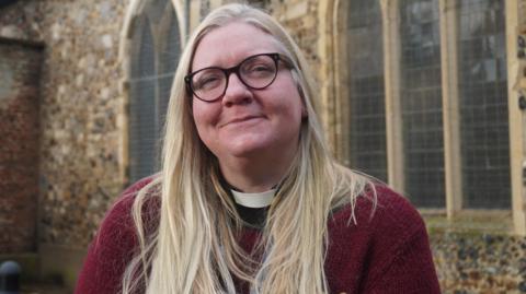 Samantha Brazier-Gibbs has long blonde hair and is wearing black rimmed glasses, a clerical collar and a burgundy jumper. She is smiling and standing in front of a flint church, with arched windows. 