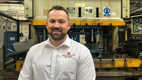 Michael Beese pictured standing in front of heavy-duty machinery. He has short black hair and a dark stubble beard. He is wearing a white collar shirt with the red Genex UK Limited logo.
