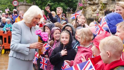 A beaming Queen Camila outside the gates of the abbey in a grey suit greeting a crown of children in colourful coats in the rain. Many are waving the Union Jack and there are adults in the background taking photos on their phones. 