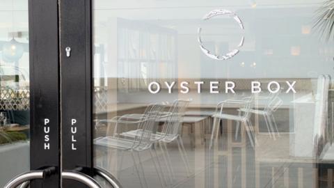 A glass door to a restaurant, with a black frame. On the glass, it says 'Oyster Box'. Above the words is a hollow white circular logo. Reflected in the glass, white metallic chairs are visible. On the black door frame are the words 'push' and 'pull' in white writing.