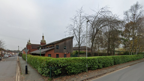 A hedge runs along the side of both streets and meets on a corner from where the photo has been taken. A red-bricked and wooden-clad building can be seen on the left of the image, behind the hedge and next to the gold-looking spires of the Ukrainian church.