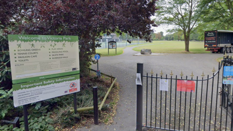 The entrance to a park. There is a black gate on the right and an opening in the path to the left of the gate for people to walk through. There is a cream and green sign that reads "Victoria Park". Beyond the gate there is a large green space and tennis court which are separated by a path.
