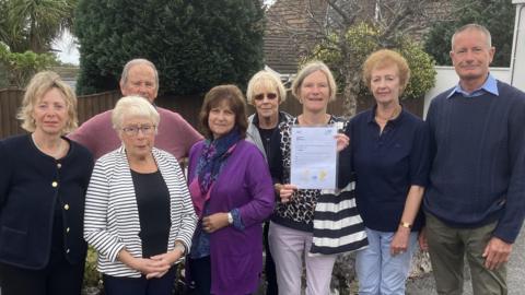 Eight people standing on a surfaced driveway in front of a low brick wall. They are all looking at the camera and one woman is holding a letter on an A4 page up but the contents of the page are too small to read