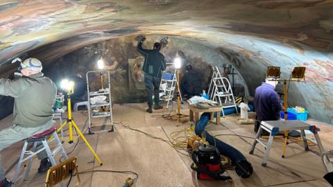 Three people, on a platformed area, in a room, with a painted ceiling above them, and painting to the end of the room. One man is standing, with an arm up, the other men are sitting down. They are all working, there are lights around them and equipment and ladders. 