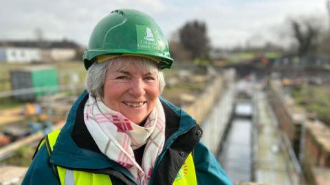 Person wearing a green hard hat and high-visibility jacket stands in front of a canal lock under construction, with building materials and equipment visible along the site