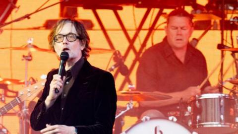 Pulp frontman Jarvis Cocker is wearing a black velvet jacket over a brown shirt and has black-rimmed glasses on. He is singing into a microphone on stage. On the right, behind him, is Nick Banks who is wearing a navy shirt and playing drums.