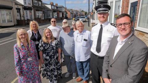 In the right is Sunderland City Council Leader Cllr Michael Mordey with Superintendent Mark Hall. From left: Deputy Leader of Sunderland City Council and Cabinet Member for Health, Wellbeing and Community Safety, Councillor Kelly Chequer, and second from left in the foreground Cabinet Member for the Environment, Councillor Lindsey Leonard with Hendon residents. They are smiling and standing on the pavement. The street behind them has rows of terraced houses and a number of parked cars.