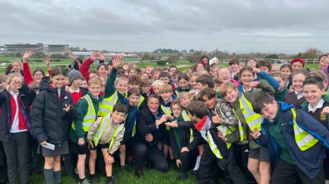 Big crowd of school students standing on Cheltenham racecourse, surrounding jockey Daryl Jacobs who has his arm in a sling. They are all smiling and cheering. Some of the children are wearing high-vis vests over their coats. 