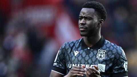 Mo Faal watches the action during a match for Wrexham. He is wearing a black and grey away shirt.