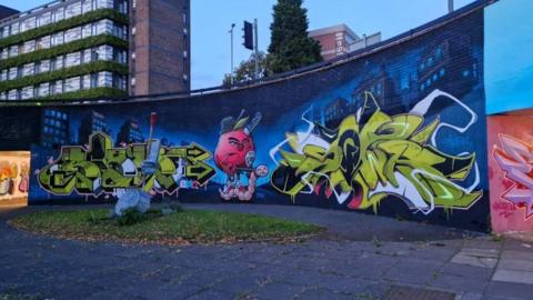 A mural is displayed on a wall in front of a paved area on a skate park on a traffic island in Penn Road. Blocks of flats are behind the wall and there are traffic lights on the road above, with a tree.