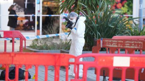 A forensic officer wearing a white suit taking photos of evidence on the ground. There are orange barricades and police tape around a train station.