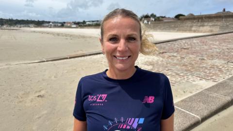 A blonde-haired woman stood on a path beside a sandy beach. 