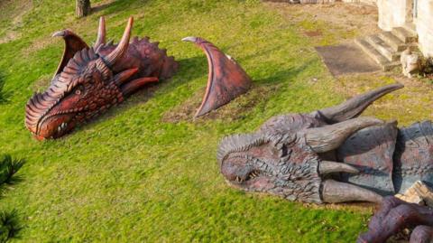Two large, scaly dragon sculpture heads are emerging out of the grass at Lincoln Castle. One is red with black bits and has part of a wing coming out of the grass and the other is grey and you can see some of its claws in the corner of the image.