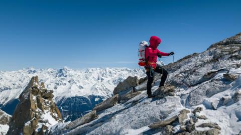 Fay Manners using ropes to reach the summit of the mountain. She is carrying equipment on her back and the sky is blue with snow covering the peaks of other mountains in the distance.