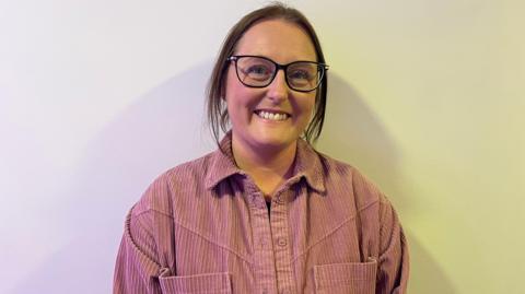a woman in a pink shirt with brown hair tied up and wearing glasses, smiling at a camera