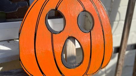 A flat piece of wood cut in the shape of a pumpkin with a face carved into it. It is painted orange