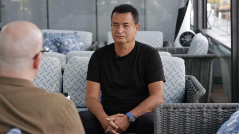A man wearing a black T-shirt sits on a sofa during an interview in a lounge area, with hands clasped together. Another person is visible out of focus in the foreground. Grey wicker furniture and patterned cushions can be seen in the background.