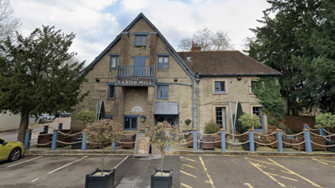 The entrance to a pub that is built from light stone. There are a five car parking spaces in front on the entrance and several bay trees. The buildings windows are painted light blue and a sign above the door reads "The Saxon Mill"