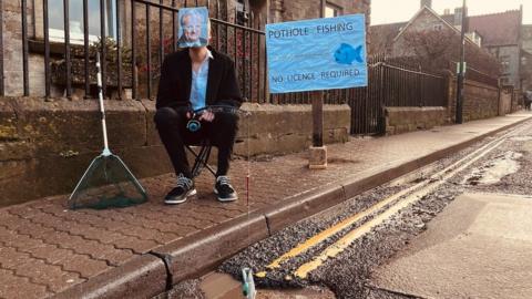 Image of Ben Thornbury. He is sat on the pavement next to a sign reading "pothole fishing no licence required". He is holding a fishing rod dangled into a water-filled pot hole in Malmesbury. His face is covered by a picture of James Gray MP.