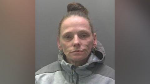 Adele Machin, a woman wearing a grey zip-up tracksuit with a hood. Her hair is scraped into a bun on the top of her head, she is wearing gold hoop earrings and looking directly at the camera.