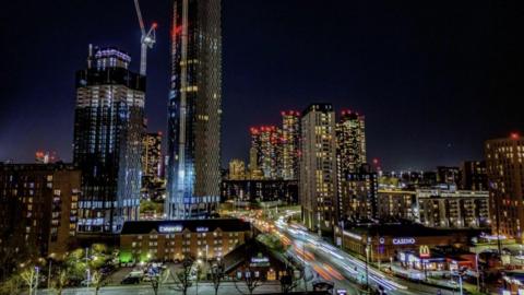 A number of high-rise buildings - making up Manchester's skyline - are in view. On the left-hand side, a crane is positioned on top of a building which is currently under construction.