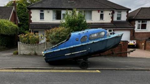 A small blue boat on the back of a trailer is seen on a pavement with a row of houses in the background.