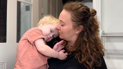 Neva Richardson in the doorway of a kitchen with her son Malin. She is holding the two-year-old in her arms, and he is leaning his head on her shoulder. Neva Richardson has long, curly red-brown hair and is wearing a black top. Malin is in a faded pink-orange T-shirt