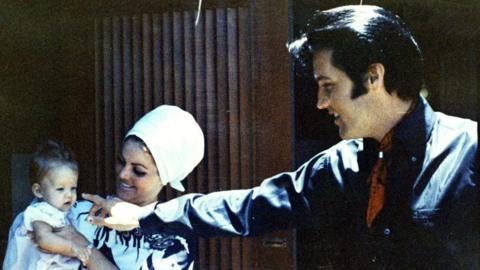 Lisa Marie Presley as a baby being held by Priscilla Presley, who is looking at her smiling and wearing a large white bonnet-style hat. Elvis Presley is standing next to them, also smiling and pointing near Lisa's face, about to touch her nose. He's wearing a black shirt with red bandana around his neck.