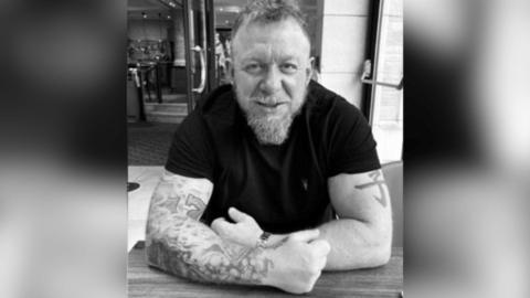 A black and white photo of Mark Killick wearing a black T-shirt and smiling at the camera, his arms are crossed on a table in front of him and his right arm is heavily tattooed, he has a beard and mustache.