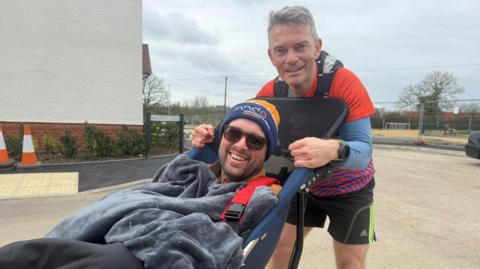 Two white men looking and smiling at the camera, one is in a wheelchair the other stands holding him.