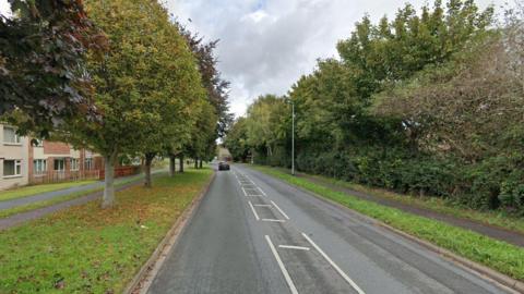 A google street view image showing a stretch of Lisieux Way, in Taunton, where the crash occurred. There are trees lining either side of the road, with pavements on either side.