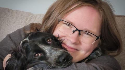 Emily Thornton-Sandy sitting on a sofa with a dog. She is hugging it to her chest and the dog had his snout on her arm. It has black fur with mottled white on the top of his nose going up between his eye. She has a broad smile and light brown hair down to her shoulders. She is wearing a grey top and glassed with a black rim on top 