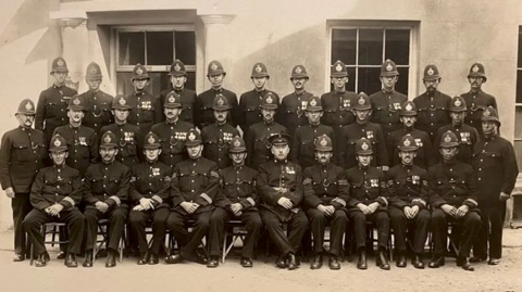 Black and white photo of Guernsey Police officers lined up in three rows for a picture. The front row is seated and the back row is stood on benches or chairs. All are in uniform with just over half of them wearing service medals. Six sergeants are seated in the front row with a senior officer in the centre.