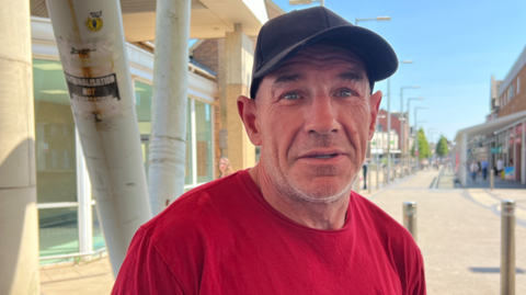 A man wearing a red t shirt and black baseball cap stood in front of a high street on a sunny day.