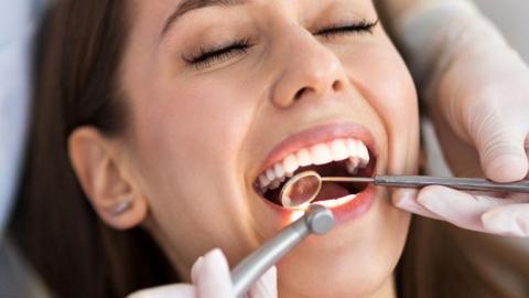 Open-mouthed dark-haired woman being examined by a dentist, who has a white jacket and is wearing gloves