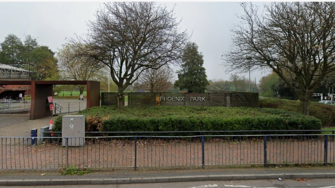 The park entrance is on a main road and can be seen behind some road-side railings. The name of the park appears on a wooden fence with the entrance either side of it and low hedge in front of it.