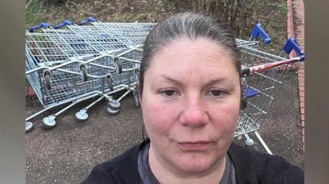 Kirsty Knight standing in front a fallen-down line of trolleys - taking a selfie- wearing a grey T-shirt and a black cardigan