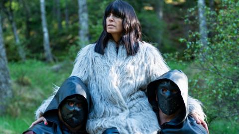 Claudia Winkleman sitting on the shoulders of two masked men in a fur coat