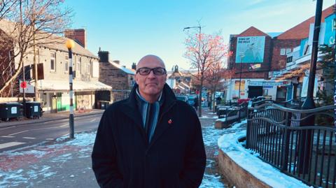 A man with a bald head and glasses is wearing a scarf and a smart winter coat. He is standing on the pavement on a high street with a few shops behind him on the right and a pedestrian crossing behind him on the left.