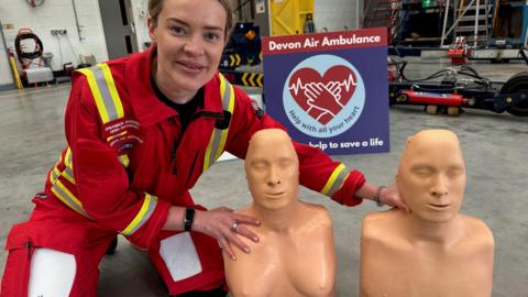 Rhiannon Roderick crouches next to two training manikins. One is a male torso and the other is a female torso. She is wearing a red paramedic jumpsuit. In the background there is a Devon Air Ambulance Sign. 