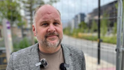 Jim Robbins looks at the camera with a serious expression. He is stood on a street in Swindon, with a metal wire mesh fence on the right, behind which blurred buildings can be seen. He has grey hair and wears a light brown t-shirt under a grey suit jacket. 