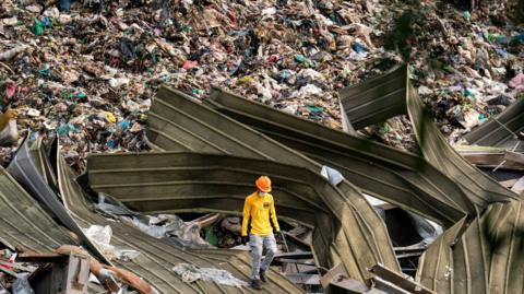Man wearing hard hat and yellow shirt and jeans walks over twisted tin roofs on the site of a rubbish landslide