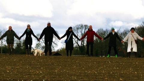 A group of people stand in a field of bare earth, holding hands. A dog is standing in front of them.