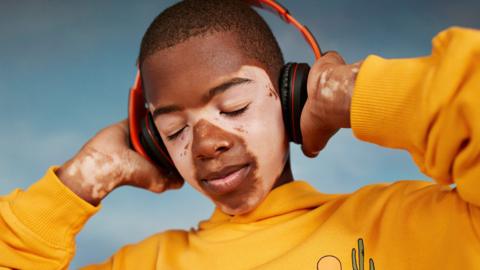A boy with a yellow hoody on wears black and red headphones. He has is eyes closed and is holding the headphones over his ears