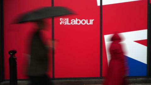 A silhouette of a delegate with an umbrella walks past a union jack sign with the Labour logo, outside the party's annual conference in Liverpool