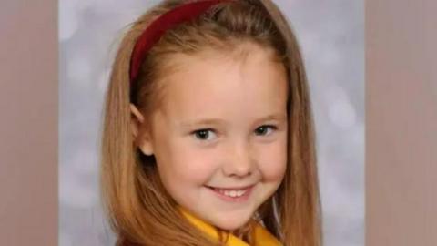 Elsie Dot Stancombe wears a red hairband and school uniform and smiles a the camera. 