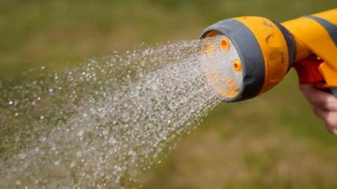 A person holding the nozzle of a yellow hosepipe with water gushing out of it. It is being sprayed onto some grass. 