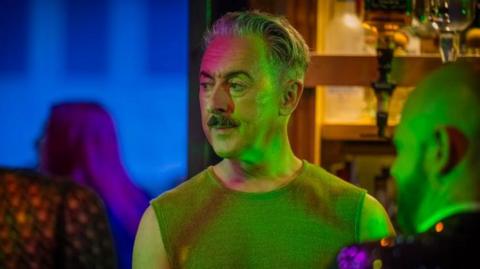 Alan Cumming is standing behind the bar during a scene from the filming of Tip Toe. Bottle