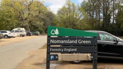 A car park. A sign in the foreground reads "Nomansland Green, Forestry England." It is black and green with a cut-in symbol of a tree on the wood. In the background several cars are parked on gravel and a road between the two sides of the car park leads into a forested area. 